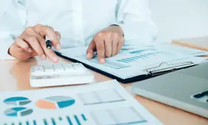 Osoba liczy na kalkulatorze, analizując wykresy i tabele. Obliczenia dotyczą raty annuitetowej.