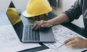 Inżynier pracuje nad planami, korzystając z laptopa i długopisu. Na biurku leży żółty kask, symbolizujący ekspertyzy budowlane.