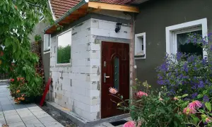 Rozbudowa budynku gospodarczego bez pozwolenia. Ściany z białych pustaków, nowe drzwi i okno. Wokół kwitnące kwiaty.