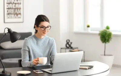 Kobieta w okularach pije kawę, pracując przy laptopie. Analizuje oferty nieruchomości, licząc prowizję biura nieruchomości.