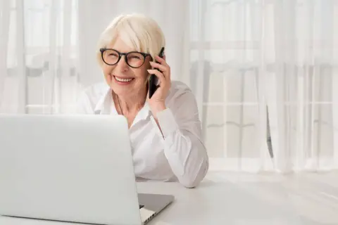 Uśmiechnięta starsza pani w okularach rozmawia przez telefon, siedząc przy laptopie. Dyskutuje o służebności mieszkania i obowiązkach właściciela.