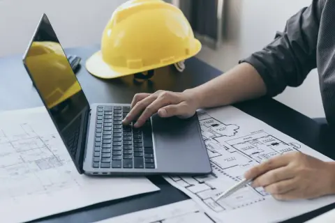 Inżynier pracuje nad planami, korzystając z laptopa i długopisu. Na biurku leży żółty kask, symbolizujący ekspertyzy budowlane.