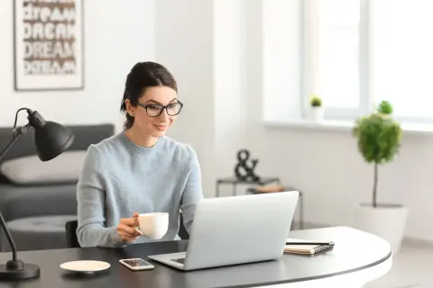 Kobieta w okularach pije kawę, pracując przy laptopie. Analizuje oferty nieruchomości, licząc prowizję biura nieruchomości.