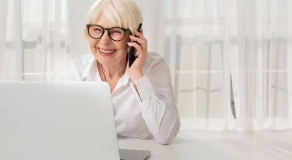 Uśmiechnięta starsza pani w okularach rozmawia przez telefon, siedząc przy laptopie. Dyskutuje o służebności mieszkania i obowiązkach właściciela.