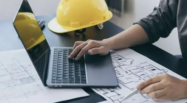 Inżynier pracuje nad planami, korzystając z laptopa i długopisu. Na biurku leży żółty kask, symbolizujący ekspertyzy budowlane.