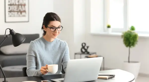 Kobieta w okularach pije kawę, pracując przy laptopie. Analizuje oferty nieruchomości, licząc prowizję biura nieruchomości.