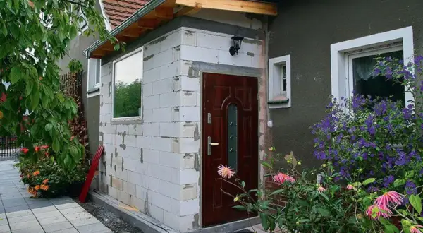 Rozbudowa budynku gospodarczego bez pozwolenia. Ściany z białych pustaków, nowe drzwi i okno. Wokół kwitnące kwiaty.