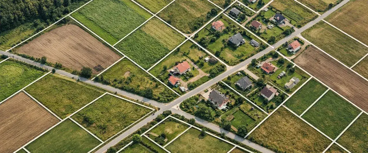 Widok z lotu ptaka na podzielone działki z r&oacute;żnymi rodzajami nieruchomości: pola uprawne, łąki i domy jednorodzinne.