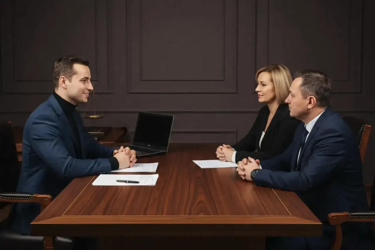 Trzy osoby przy drewnianym stole omawiają sprzedaż mieszkania za got&oacute;wkę. Na stole dokumenty i laptop.