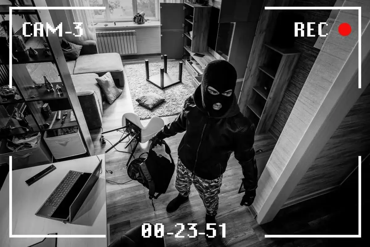 Zapis monitoringu: włamanie do mieszkania w bloku. Złodziej w masce na twarzy, z torbą w ręku, przeszukuje pok&oacute;j.