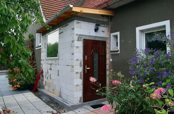 Rozbudowa budynku gospodarczego bez pozwolenia. Ściany z białych pustaków, nowe drzwi i okno. Wokół kwitnące kwiaty.